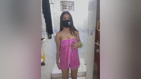 Media: Video of an Asian woman with long black hair, wearing a pink towel, black mask, and green scrunchie, standing in a small, cluttered bathroom.