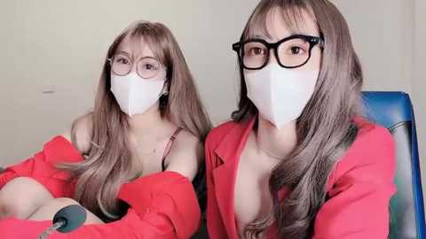 Media: Video of two women with long brown hair, wearing black-framed glasses, white face masks, and red blazers, sitting in chairs, one holding a microphone, in a light-colored room.