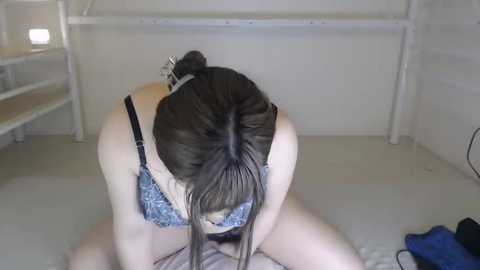 Media: A video shows a young woman with long brown hair in a bun, wearing a blue floral-patterned bra, kneeling on a bed with a white wall and metal bedframe behind her.