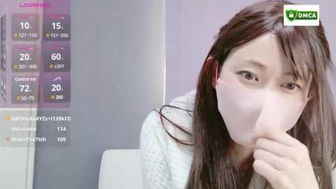 Media: Video of an Asian woman in a white mask, covering her mouth, wearing a white sweater, with a blurred indoor background and digital health tracking interface overlaid.