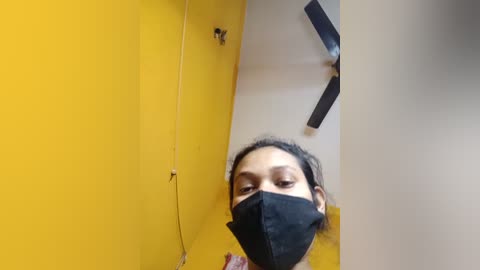 Media: Video of a woman with medium brown skin and dark hair, wearing a black face mask, standing in a yellow-walled room with a ceiling fan, and a partially visible clock.