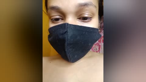 Media: Video of a young person with light brown skin, wearing a black face mask and a red-patterned shirt, partially visible in the blurred background.