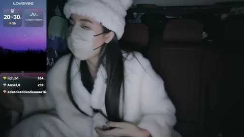 Media: A dimly lit video captures a young Asian woman with long black hair, wearing a white furry coat and a matching hat, sitting in a car. She has a mask on and appears to be playing a video game.