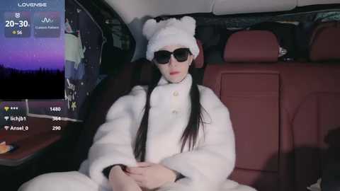 Media: Video of a woman in a white fur coat, black sunglasses, and bear hat, sitting in a car. Nighttime cityscape visible through the window, with a digital display showing location, time, and weather.