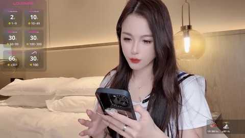 Media: A video of an East Asian woman with long, straight black hair, wearing a white blouse and black vest, holding a smartphone with a weather app displaying rain and temperature. She's in a modern, minimalist bedroom with beige walls and a cozy bed.