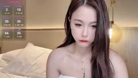 Media: Video of a young Asian woman with long black hair, wearing a white strapless top, sitting on a bed in a dimly lit bedroom.