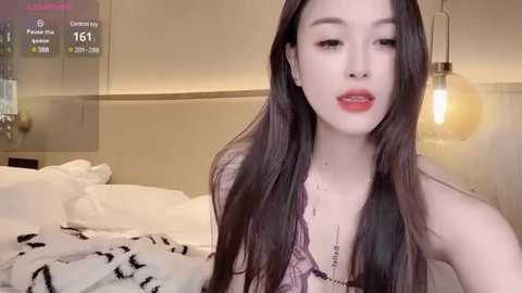 Media: Video of a young Asian woman with long black hair, fair skin, and red lipstick, wearing a purple lace bra, in a modern, warmly lit bedroom with white bedding, a glass table, and a hanging light bulb.