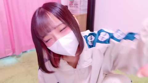 Media: A video of an Asian woman with straight, shoulder-length dark hair, wearing a white face mask and a white hoodie with blue patches, sitting on a light green carpet. The background features pink curtains and a wooden shelf with papers.