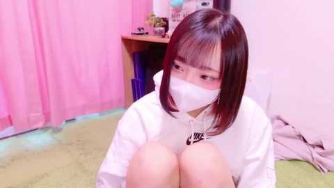 Media: Video of an Asian woman with straight, shoulder-length brown hair, wearing a white Nike hoodie, face mask, and sitting on a green carpet. Background features pink curtains, a wooden desk, and scattered items.