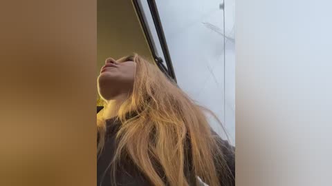 A video captures a young woman with long, light brown hair, tilted head, and closed eyes, standing near a window with a blurred, cloudy sky outside.