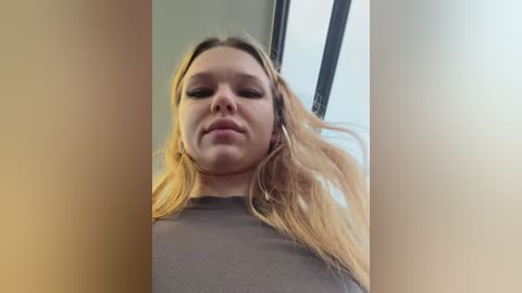 Video of a young woman with long blonde hair, wearing a dark shirt, captured from a low angle, indoors with blurred background.