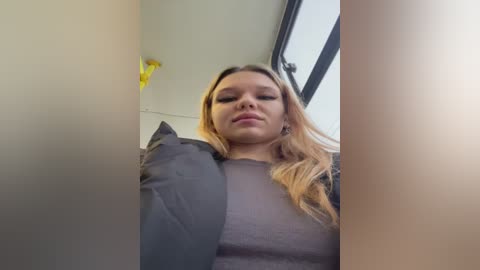 Media: A video of a young woman with long, blonde hair, wearing a gray sweater, captured from a low angle, with blurred background showing a yellow object and a window.