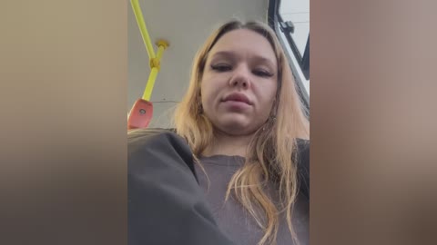 Media: Video of a young Caucasian woman with long blonde hair, wearing a black sweater, looking straight ahead. Background shows a public bus interior with yellow poles and a red sign.