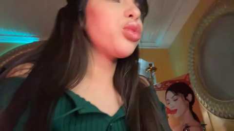 Media: Video of a woman with long black hair and fair skin, wearing a green blouse, puckering her lips in a kiss. Background features a golden-framed mirror and a portrait.