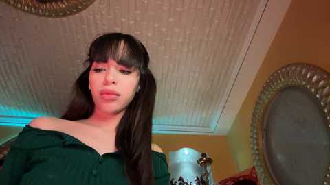 Media: Video of a young woman with long, dark hair and a pale complexion, wearing an off-shoulder green top. Background features a textured ceiling, a round mirror, and a lamp.