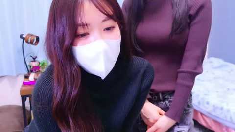 Media: Video of two Asian women in a bedroom; one with long dark hair, white face mask, and black sweater, the other in a dark turtleneck, assisting with a mask.