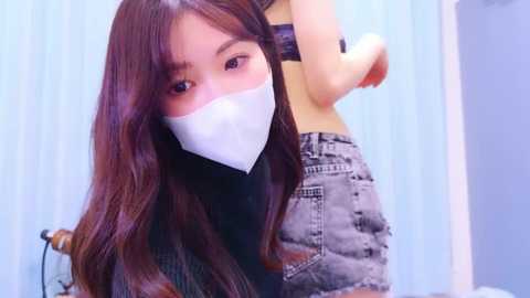 Media: A video of an East Asian woman with long, straight brown hair, wearing a white surgical mask and a black top, standing in a room with light blue vertical blinds.