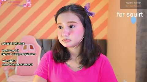 Media: Video of a young woman with medium-length black hair, wearing a pink shirt, seated indoors against a striped orange and yellow wall. Text overlay includes \"LoveSINMASS\" and \"Every for gummy pleasure.\