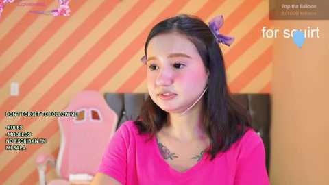 A video of a young Asian woman with straight black hair and a purple hair clip, wearing a pink shirt. She's seated in a room with orange-striped walls and a pink gaming chair.