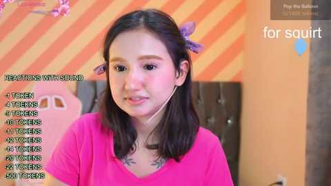 Media: Video of a young Asian woman with shoulder-length black hair, wearing a pink shirt, speaking into a microphone. Background includes a striped wall and a pink flower.