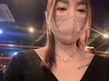 Media: Video of an Asian woman with long, pink-streaked hair wearing a black V-neck top and a clear face mask. She has a small tattoo on her left shoulder and is standing indoors with blurred stadium lights in the background.