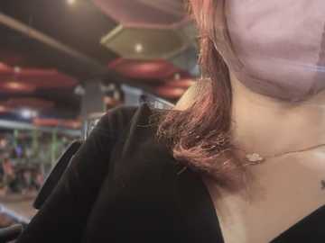 Media: Video of a woman with long, wavy brown hair, wearing a light pink top and black jacket, seen from the side in a modern, dimly-lit indoor setting with blurred background.