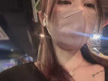 Media: Video of an Asian woman with light skin, wearing a surgical mask, black top, and a green headband, with a tattoo on her right shoulder. Background is blurred, suggesting an indoor setting.