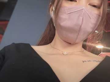 Media: Video of a woman with light skin, brown hair, wearing a pink surgical mask and black top, with a small tattoo on her neck.