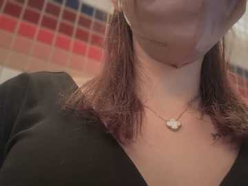 Media: A video of a woman with light skin, wearing a black V-neck top and a facemask, her shoulder-length hair styled with a fringe. Background shows a tiled wall with red and blue squares.