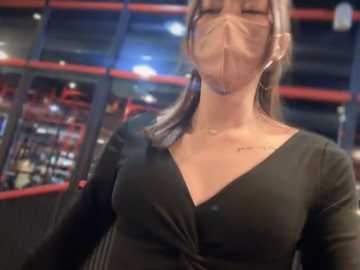 Media: Video of an Asian woman with medium-length, straight, light brown hair, wearing a black V-neck top, a clear face mask, and a black crossbody bag, indoors with dim lighting, blurred bookshelves in the background.