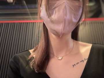 Media: Video of a woman with long brown hair wearing a face mask, black top, and a necklace with a small pendant. Background shows blurred, dark interior with ribbed panels.