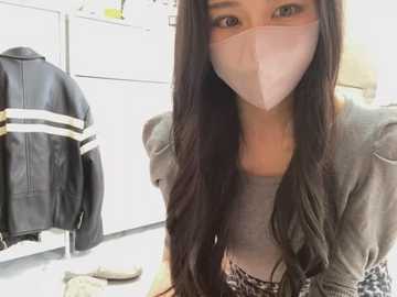 Media: Video of an East Asian woman with long, wavy black hair, wearing a gray jacket and a face mask. She kneels in a modern, white-walled laundry room with a black leather jacket hanging on a rack.