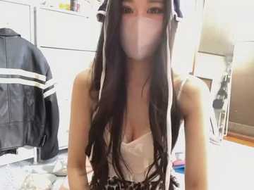 Media: A video of an Asian woman with long, straight black hair and a white surgical mask, wearing a low-cut white top, sitting in a cluttered room with a black jacket, white dresser, and a mirror.