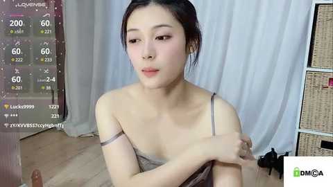 Media: A video of an East Asian woman with fair skin and straight black hair, wearing a gray spaghetti-strap dress, sitting on a wooden floor, with a white curtain background, and a \"DMCA\" watermark.