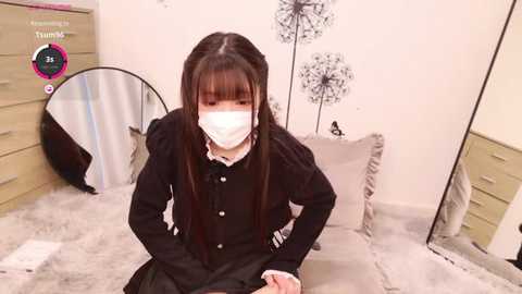 Media: Video of a young East Asian woman with long black hair, wearing a black cardigan, white mask, and skirt, sitting on a beige bed in a minimalist bedroom with white walls, a large mirror, and a white dresser.