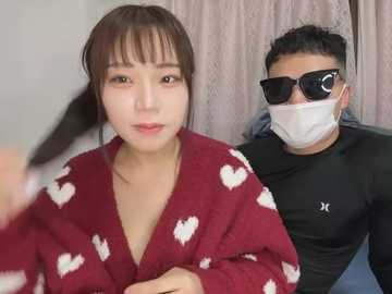 Media: Video of an East Asian woman with fair skin, brown hair, and a maroon robe with white heart patterns, standing next to a man wearing black sunglasses, mask, and a black shirt, both indoors.