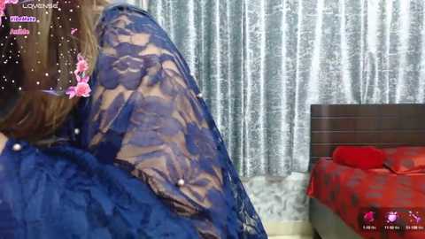 Media: Video of a woman in a blue lace dress, seated in a room with a red-patterned bedspread, white curtains, and pink flower decoration.