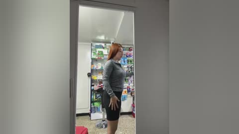 Video of a woman with long red hair, wearing a grey shirt and black skirt, standing in a brightly lit pharmacy, surrounded by colorful shelves of products.