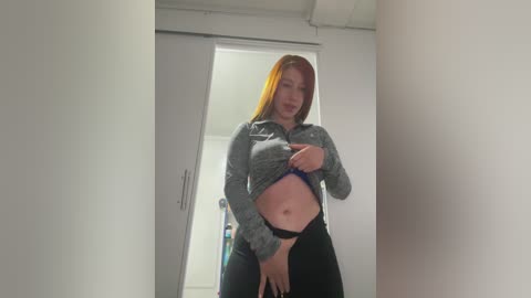 Media: A video of a red-haired woman lifting her gray sweatshirt, revealing her black bra and black pants, standing in a white hallway with closed doors in the background.