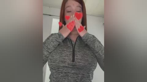 Media: Video of a fair-skinned woman with long, straight red hair, wearing a grey zip-up hoodie, holding her hands in front of her face, covered in red heart stickers, in a blurred, indoor setting.