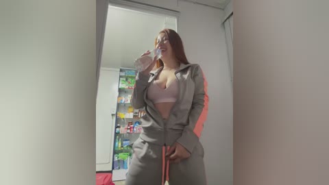 Video of a young woman with fair skin, wearing a gray tracksuit with a pink bra visible, holding a bottle. She stands in a brightly lit, modern room with a refrigerator in the background.