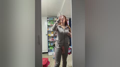 Media: Video of a young woman in a gray tracksuit, drinking from a water bottle in a modern, brightly lit kitchen with open cabinets and a messy countertop.