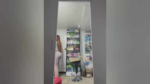 Media: Video of a cluttered, brightly-lit kitchen with a woman in a pink sweater, standing near a messy counter filled with colorful bottles, shelves, and a red stool.