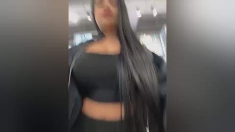 Media: A video of a woman with long black hair and medium skin tone wearing a black crop top and jacket, standing in a modern, brightly lit indoor space with blurred background.