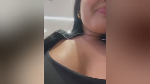 Media: A video featuring a close-up of a woman with medium skin tone and dark hair, wearing a black top, slightly open, revealing her cleavage. The background is blurred, with a hint of a white ceiling.