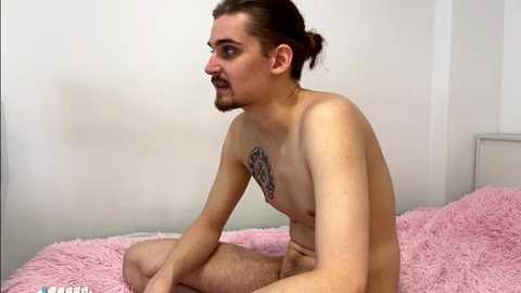 Media: Video of a nude, slender, fair-skinned man with long brown hair in a bun, sitting on a pink, fluffy bed in a minimalistic, white room.