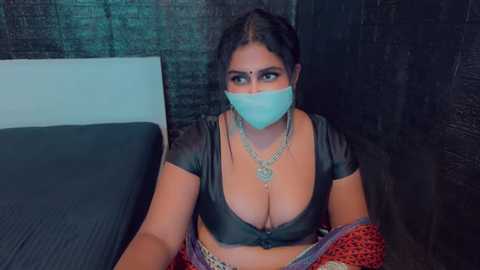 Video of a South Asian woman with dark skin, wearing a black crop top that reveals ample cleavage, a face mask, and a colorful saree, sitting on a bed with a dark wall in the background.