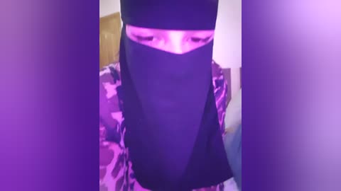 Media: A video of a person wearing a black niqab with a pinkish-purple light emanating from the eye slit, revealing only their eyes. The background is indistinct, with blurred colors.