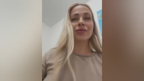 Media: A video of a blonde woman with long, straight hair, wearing a beige top, standing in a narrow, beige hallway with a blurred blue and green painting on the wall.