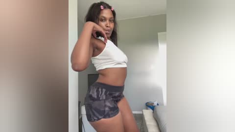 Media: A video of a young Black woman with medium-dark skin and long, straight hair styled with pink bows. She wears a white crop top and dark patterned shorts, standing indoors in a minimalist room with light walls and a beige couch.